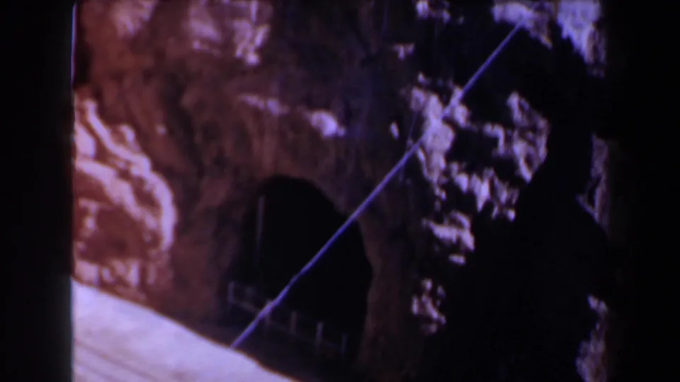 1958 Texas home-movie frame with a dark tunnel mouth in reddish rock, crossed by a diagonal cable or wire and bordered by heavy shadow.
