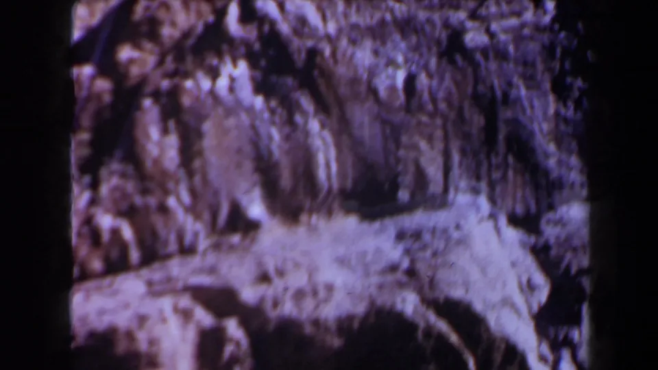 A grainy 1958 Texas home-movie frame shows uneven cliff texture, a pale stone shelf, and deep shadows across the rock.