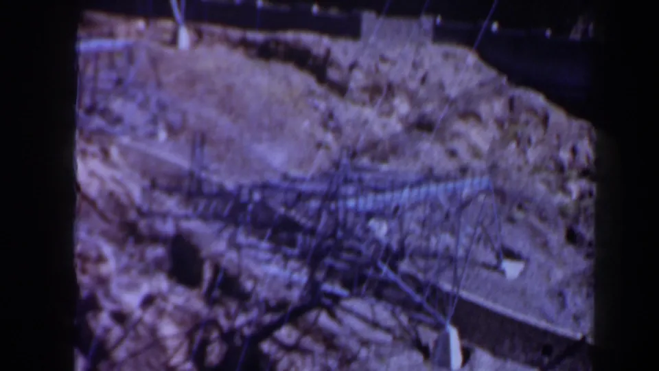 1958 Texas 8mm frame showing a rocky cliff face, stretched wires, dark recesses, and rough dirt terrain.