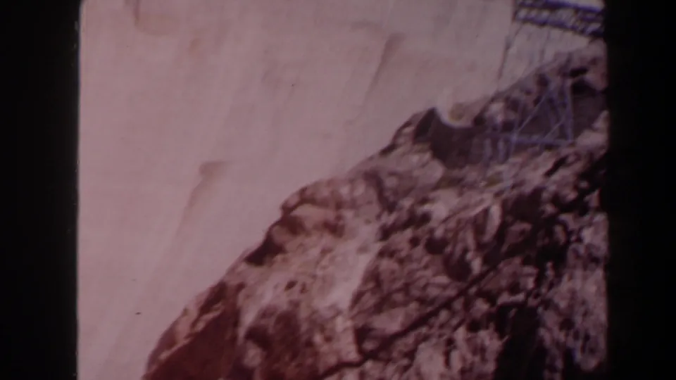 1958 Texas 8mm view of a sloping dam surface, adjacent rock, and part of a metal transmission structure near the frame edge.