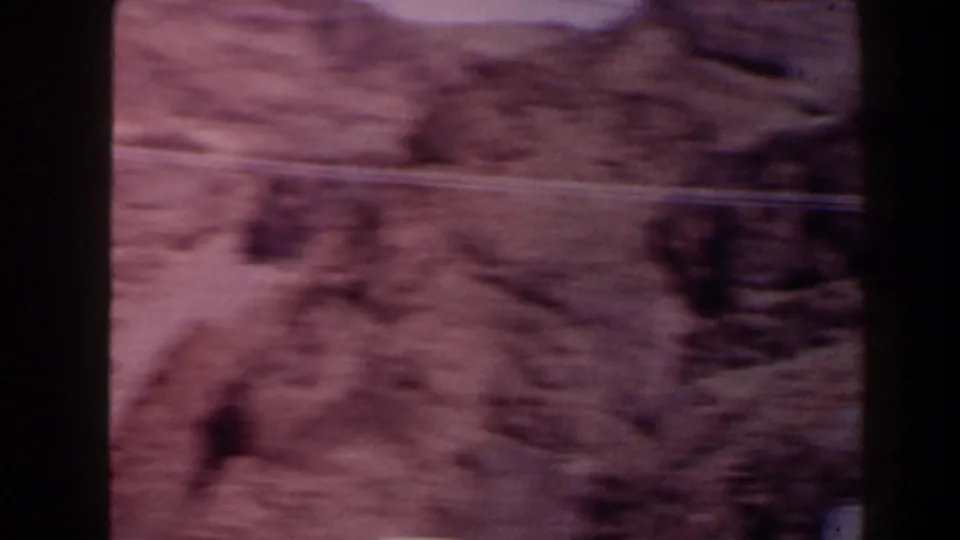 Motion-blurred 1958 Texas 8mm view of dry canyon ground, rocky slopes, and faint road or utility lines crossing the scene.