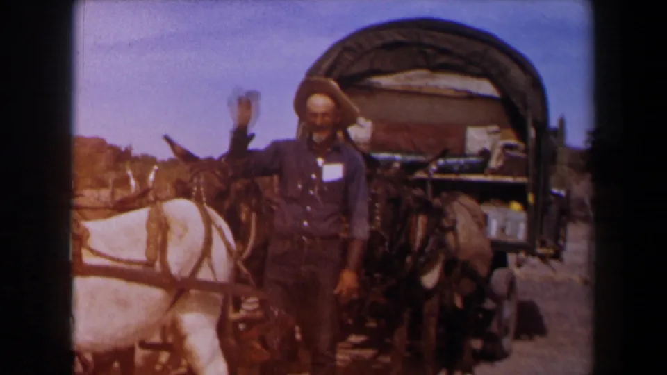 1958 Texas frame of a man in cowboy hat waving beside a covered wagon hitched to mules under bright sky.