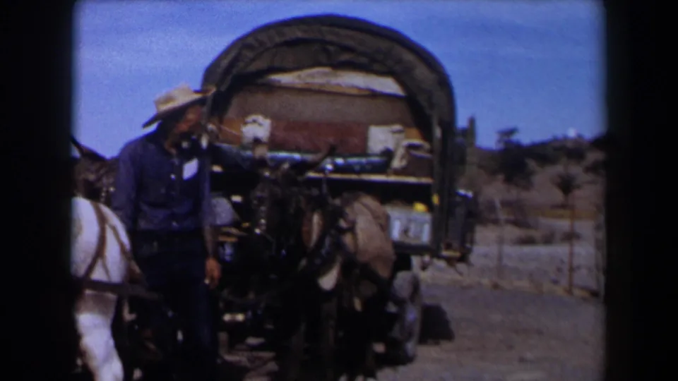 1958 Texas frame showing the man beside the mule team and covered wagon, turned away from the camera in a dry open area.