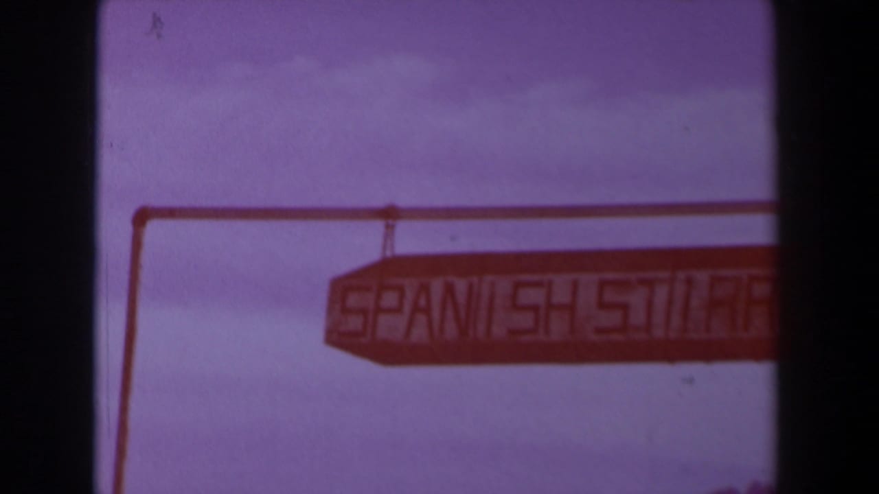 1958 Texas 8mm frame showing a wooden Spanish Street sign suspended from a horizontal support against a pale sky.
