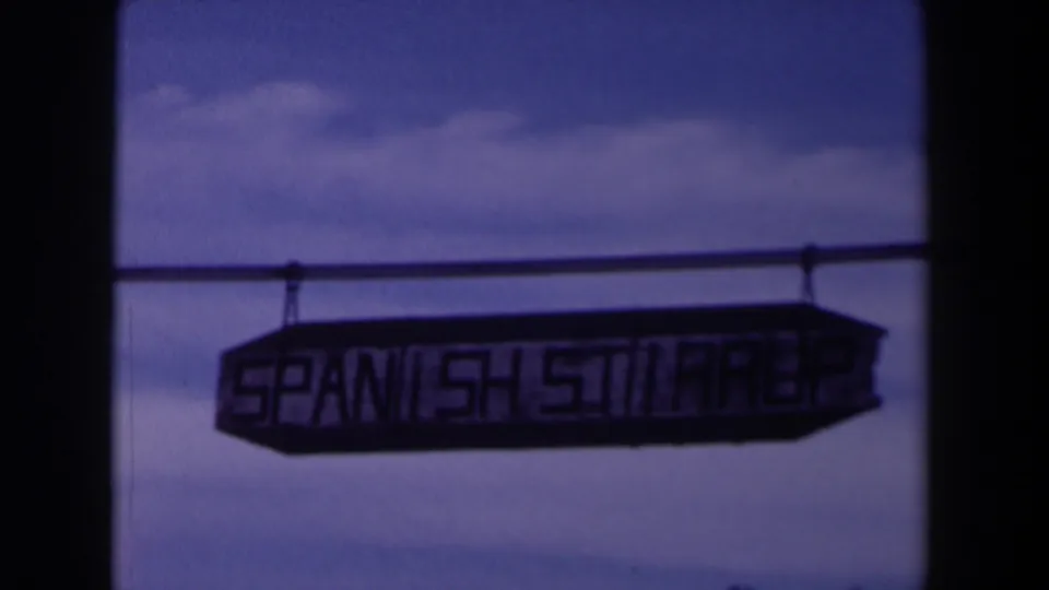1958 Texas 8mm frame showing the Spanish Street sign darker in exposure beneath a rail against a blue, clouded sky.