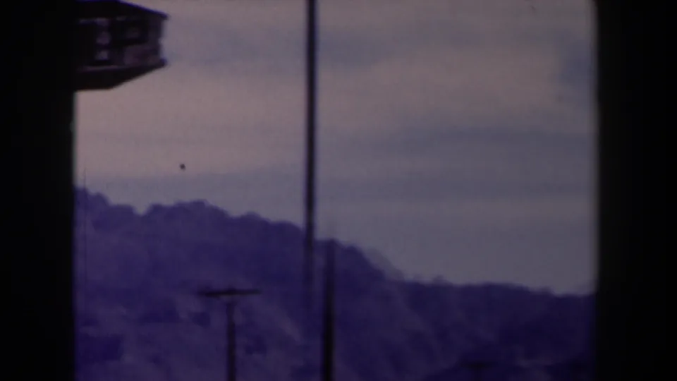 1958 Texas 8mm landscape frame with blue mountains, pale sky, vertical poles, and part of a hanging sign at the left edge.