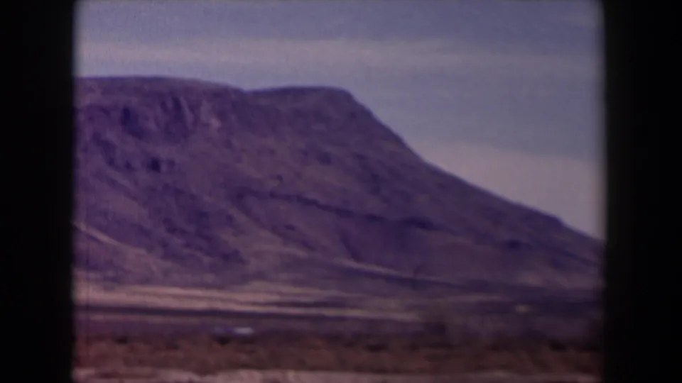 A large rocky mountain in the Texas landscape appears beyond arid ground, partially framed by the dark edges of a vehicle window.