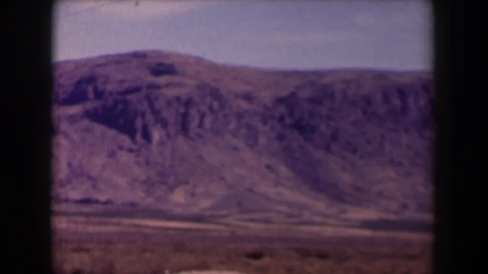 A rounded rocky hill in the 1958 Texas landscape is seen through the vehicle-window frame, with dry foreground and soft home-movie texture.