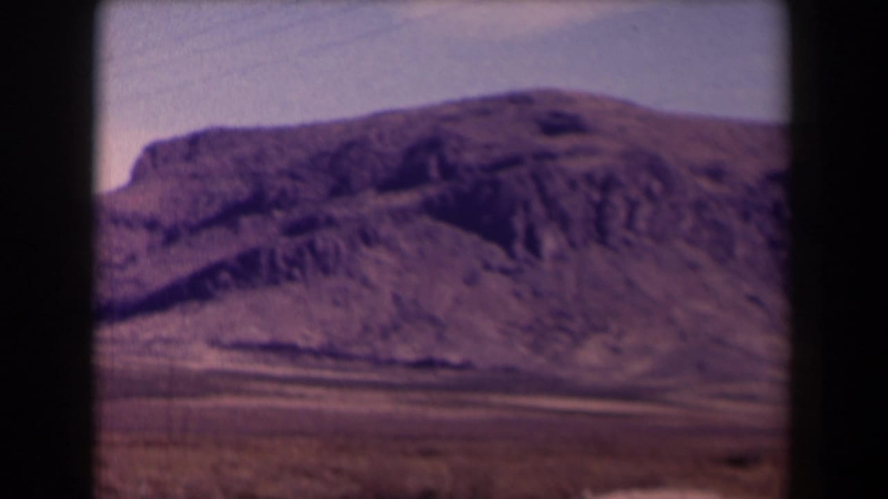 1958 Texas 8mm frame showing a dry rocky mountain slope, sparse scrub, and pale sky from a roadside travel viewpoint.