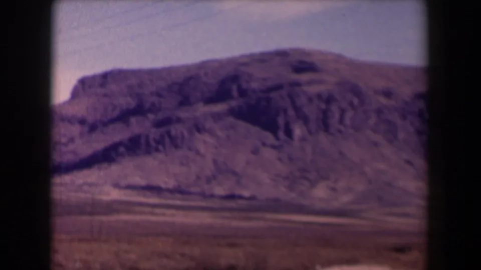 1958 Texas 8mm frame showing a dry rocky mountain slope, sparse scrub, and pale sky from a roadside travel viewpoint.