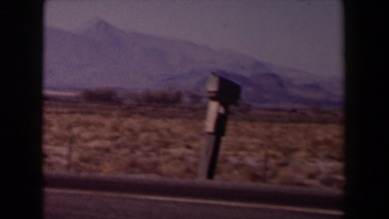 Desert roadside with a mailbox-like object, sparse brush, and distant mountains.