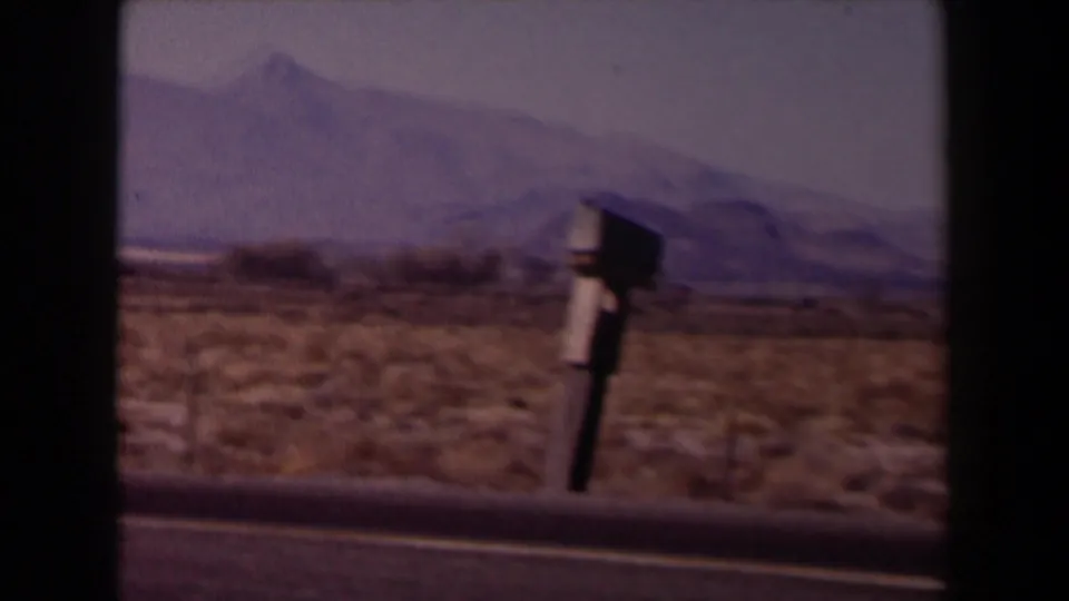 1958 Texas 8mm frame showing a desert roadside, a mailbox-like object, low brush, pale sky, and distant mountain ridges.