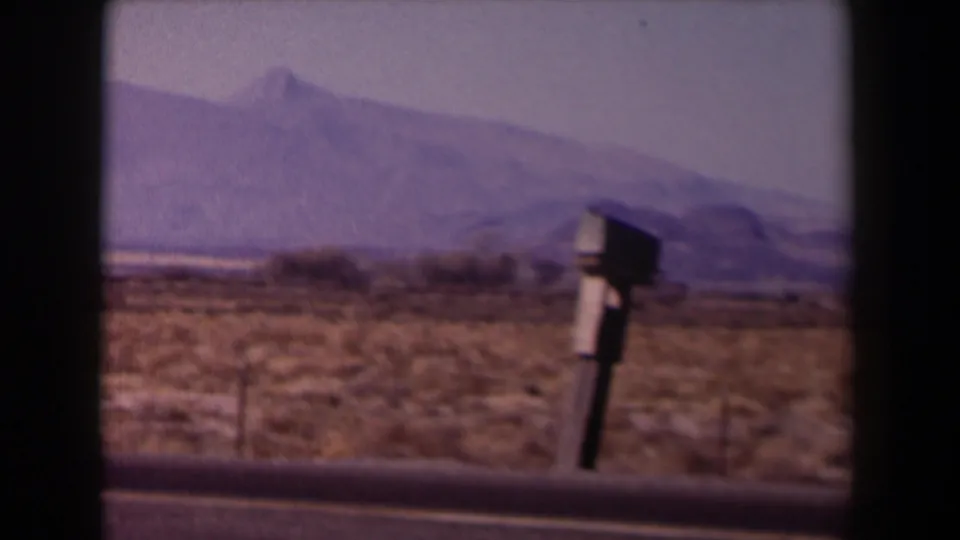 1958 Texas travel frame with a roadside mailbox-like object, open desert brush, road edge, and hazy mountain backdrop.