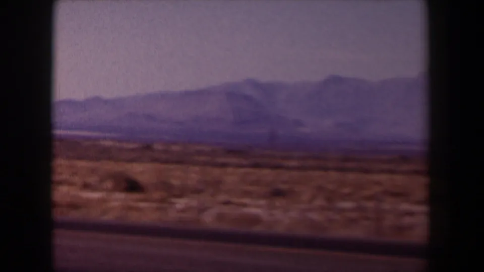 1958 Texas home-movie frame with a dark road edge, dry desert brush, pale sky, and distant mountain ridges.