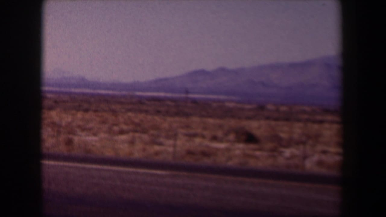 1958 Texas home-movie frame showing roadside scrubland, a paved highway edge, hazy sky, and distant low mountains.
