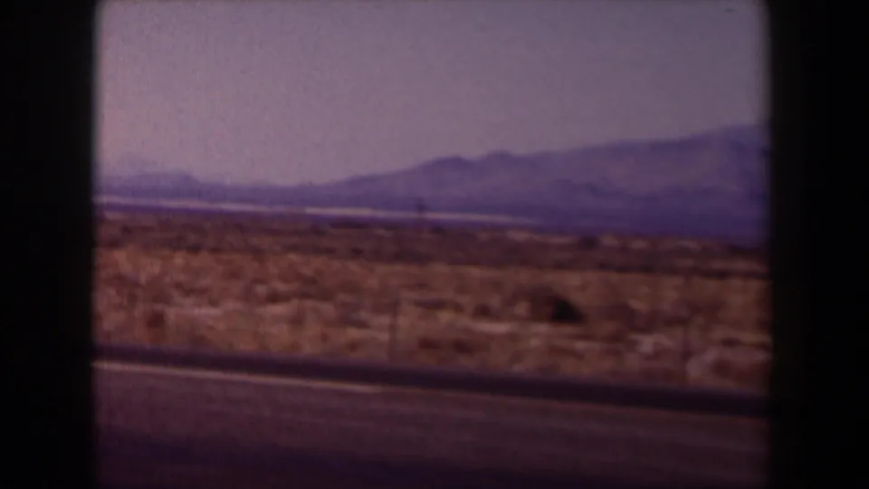 1958 Texas home-movie frame showing roadside scrubland, a paved highway edge, hazy sky, and distant low mountains.