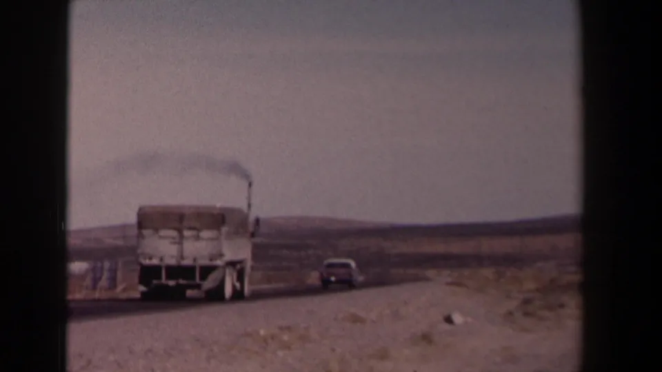 1958 Texas home-movie frame showing a white cargo truck and grey car on a highway, with low hills and open roadside land.