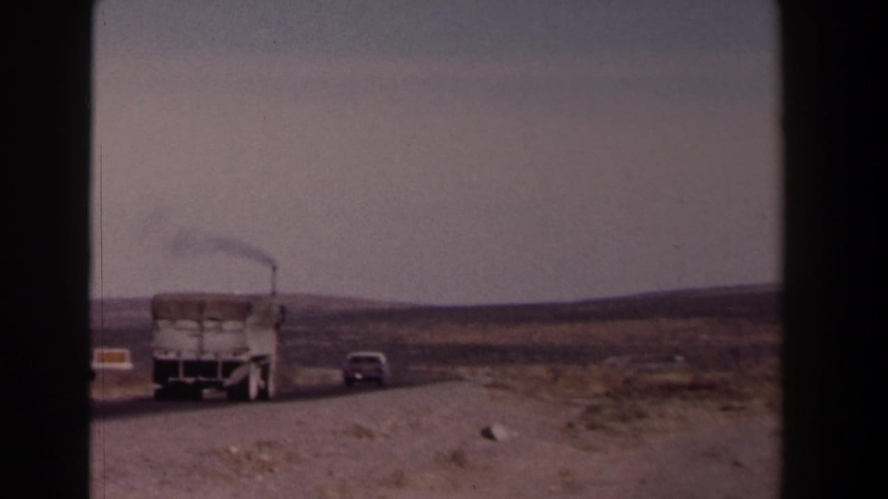 White truck follows a gray car on a dusty rural road under a hazy sky.
