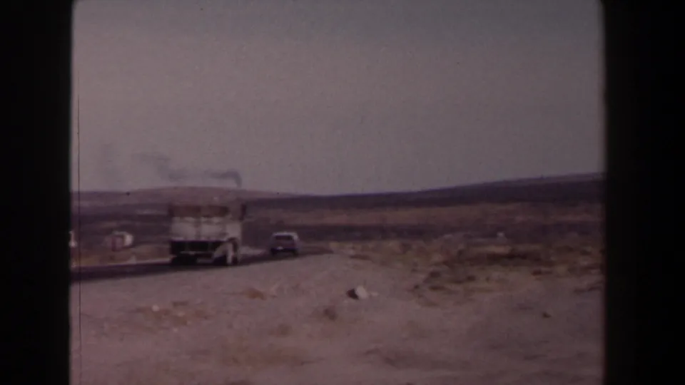 1958 Texas road footage frame with a white truck following a gray car across a dry, open roadside landscape.