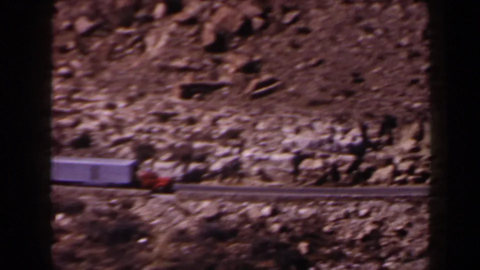 1958 Texas road scene with a red-and-gray big rig traveling past rocky terrain in soft, grainy 8mm color.