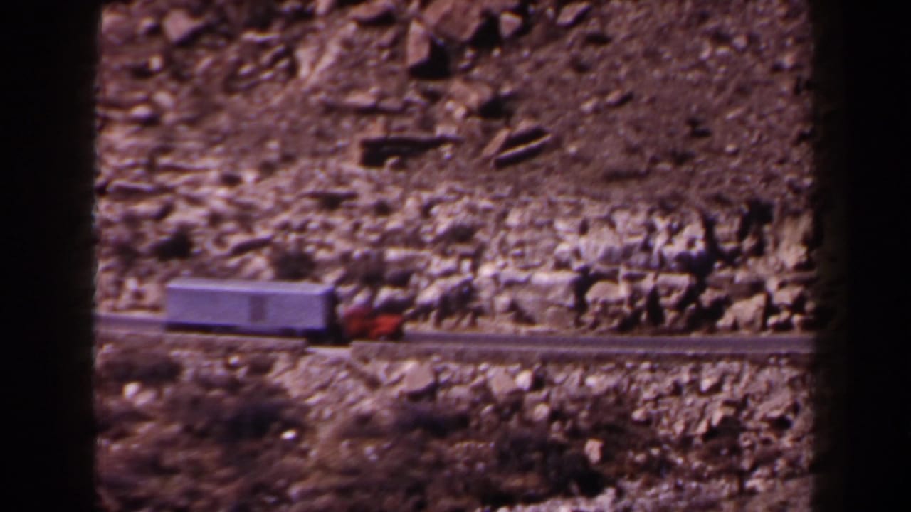 1958 Texas 8mm frame showing a red truck cab pulling a white trailer along a narrow rocky mountain road.