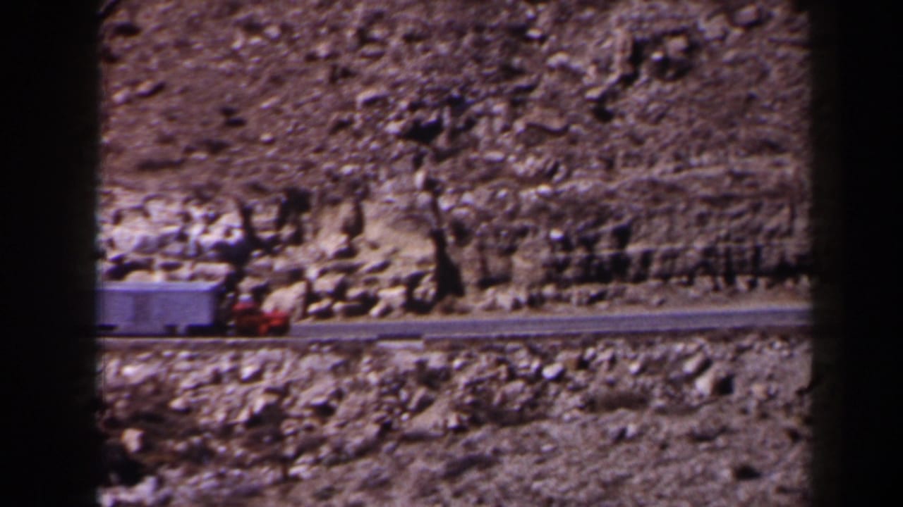 1958 Texas 8mm frame showing a red cab and white trailer traveling along a narrow road beside a rocky mountainside.