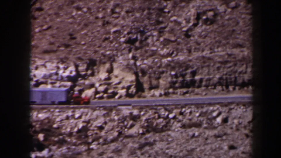 1958 Texas 8mm frame showing a red cab and white trailer traveling along a narrow road beside a rocky mountainside.