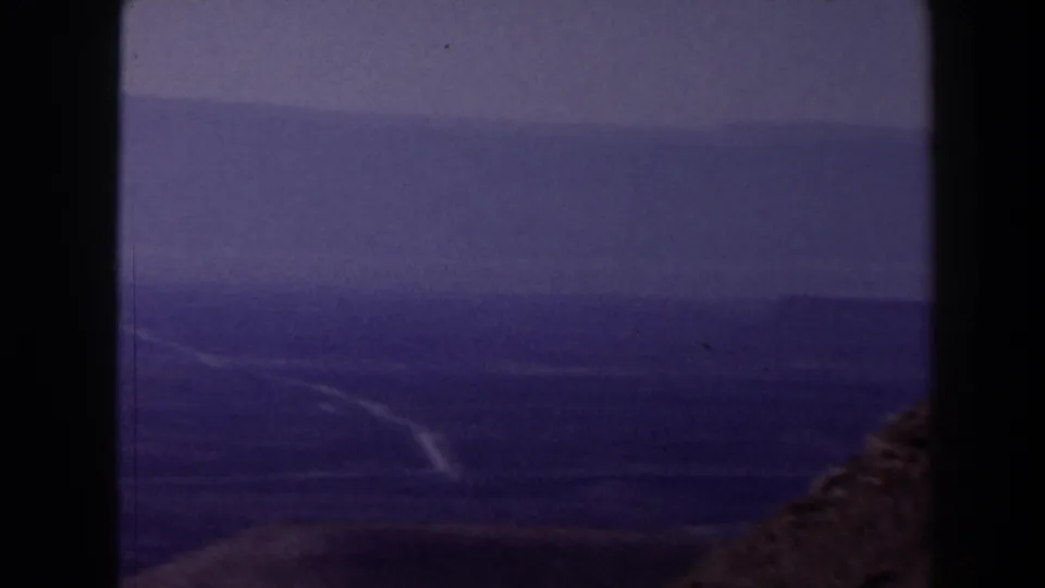1958 Texas home-movie frame with a distant valley, winding pale path, brown slope at right, and overcast sky.