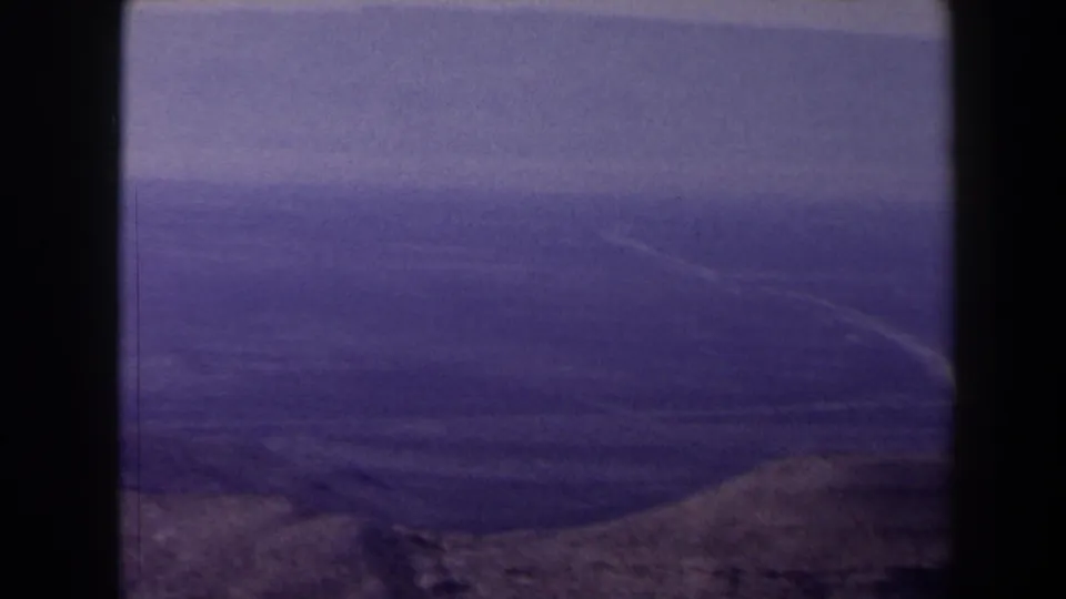Aged 8mm view of Texas fields and distant ridges, with a winding pale path and brown foreground slope under cloud cover.