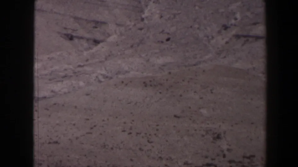 1958 Texas frame showing an arid elevated slope with scattered rocks, faint mountain forms, and subdued 8mm color.