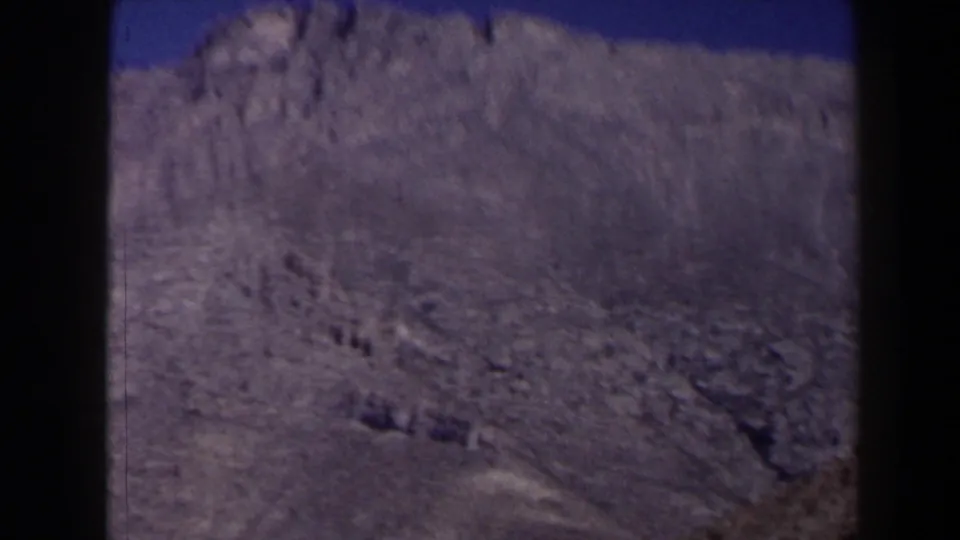 1958 Texas 8mm frame of a broad pale cliff face with rugged texture and sparse arid ground.