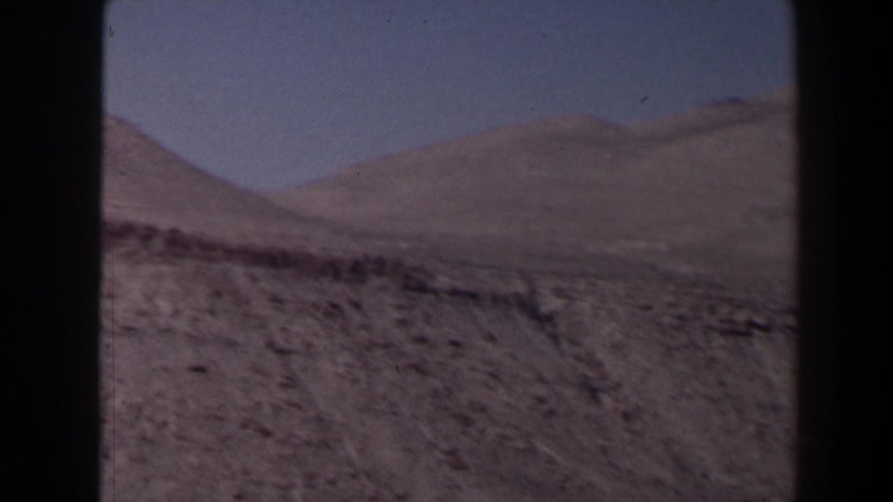 1958 Texas 8mm view across rugged canyon terrain toward rounded mountain ridges beneath a clear blue sky.