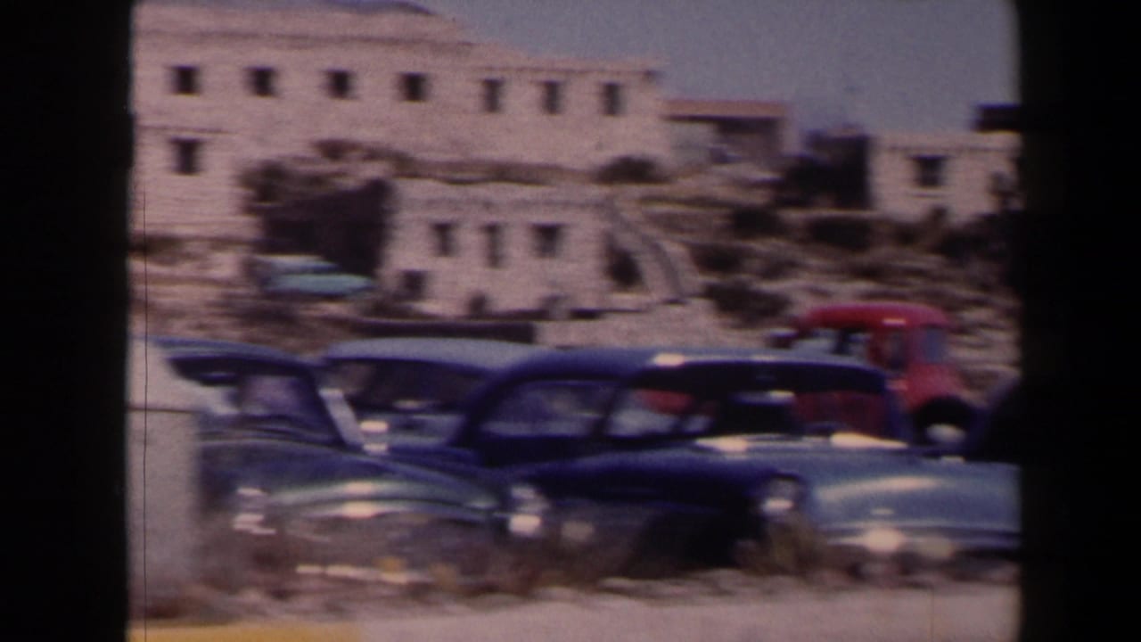 1958 Texas frame with several parked American cars below pale adobe-style buildings in bright desert light.