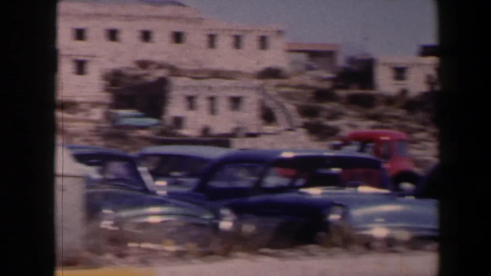 1958 Texas frame with several parked American cars below pale adobe-style buildings in bright desert light.
