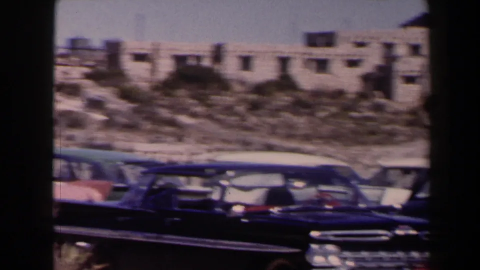 1958 Texas home-movie frame showing dark and light mid-century cars parked beneath low desert buildings.