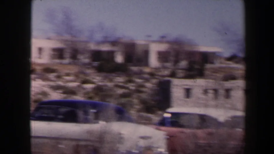 1958 Texas frame with parked cars, rocky desert slope, and pale adobe-style buildings in soft 8mm color.