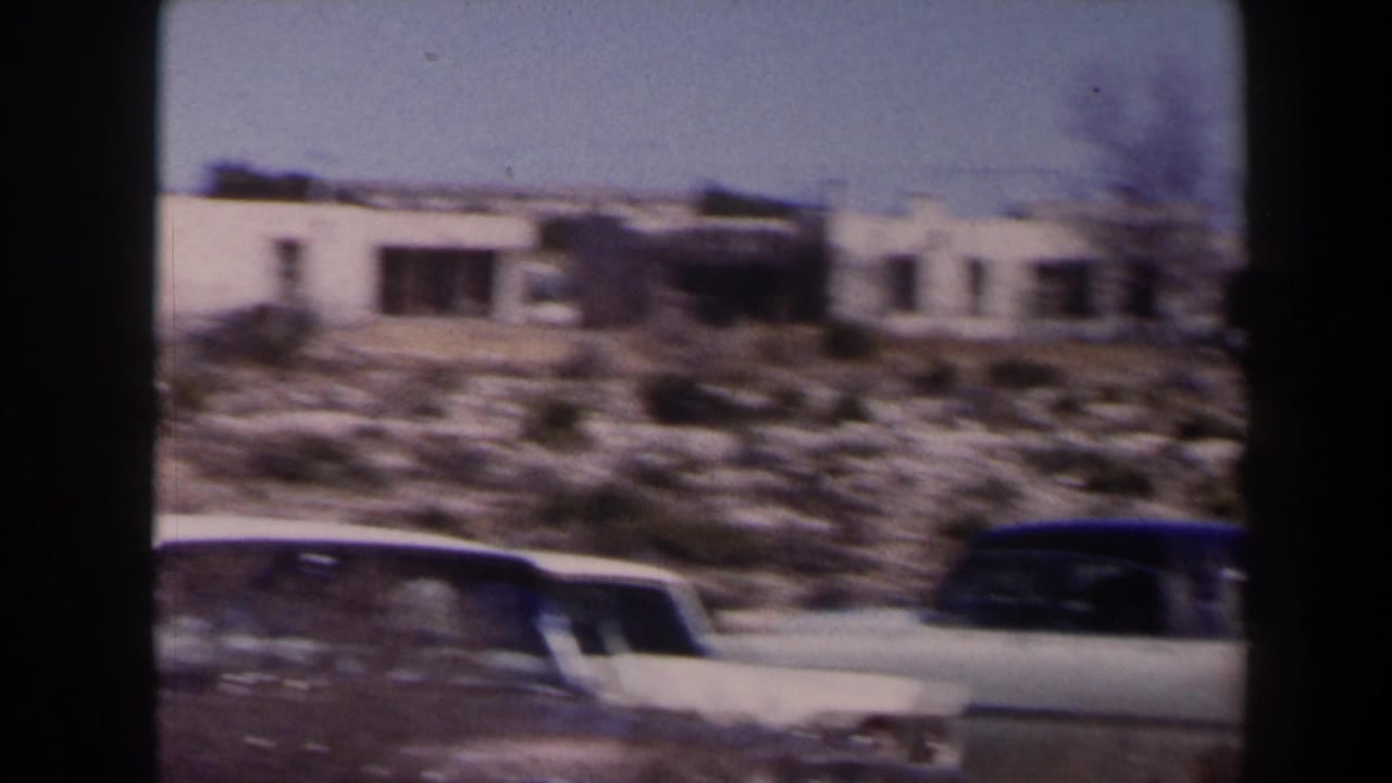 1958 Texas home-movie frame showing parked mid-century cars outside low pale buildings along a sunlit dry roadside.