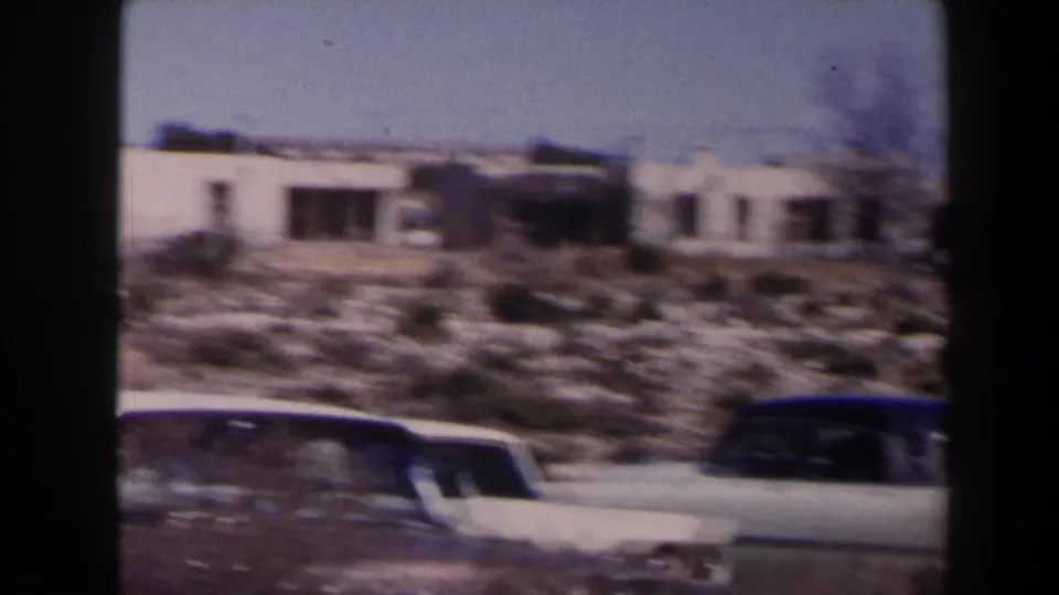 1958 Texas home-movie frame showing parked mid-century cars outside low pale buildings along a sunlit dry roadside.