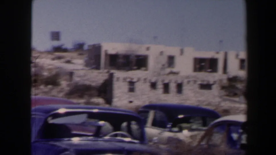 1958 Texas frame with multiple parked mid-century automobiles, low storefront-like buildings, dry brush, and film-frame borders.