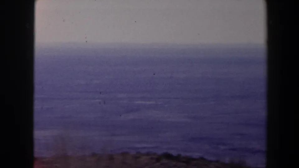 1958 Texas 8mm seascape with calm water, a pale sky, dark window framing, and faint foliage near the lower edge.
