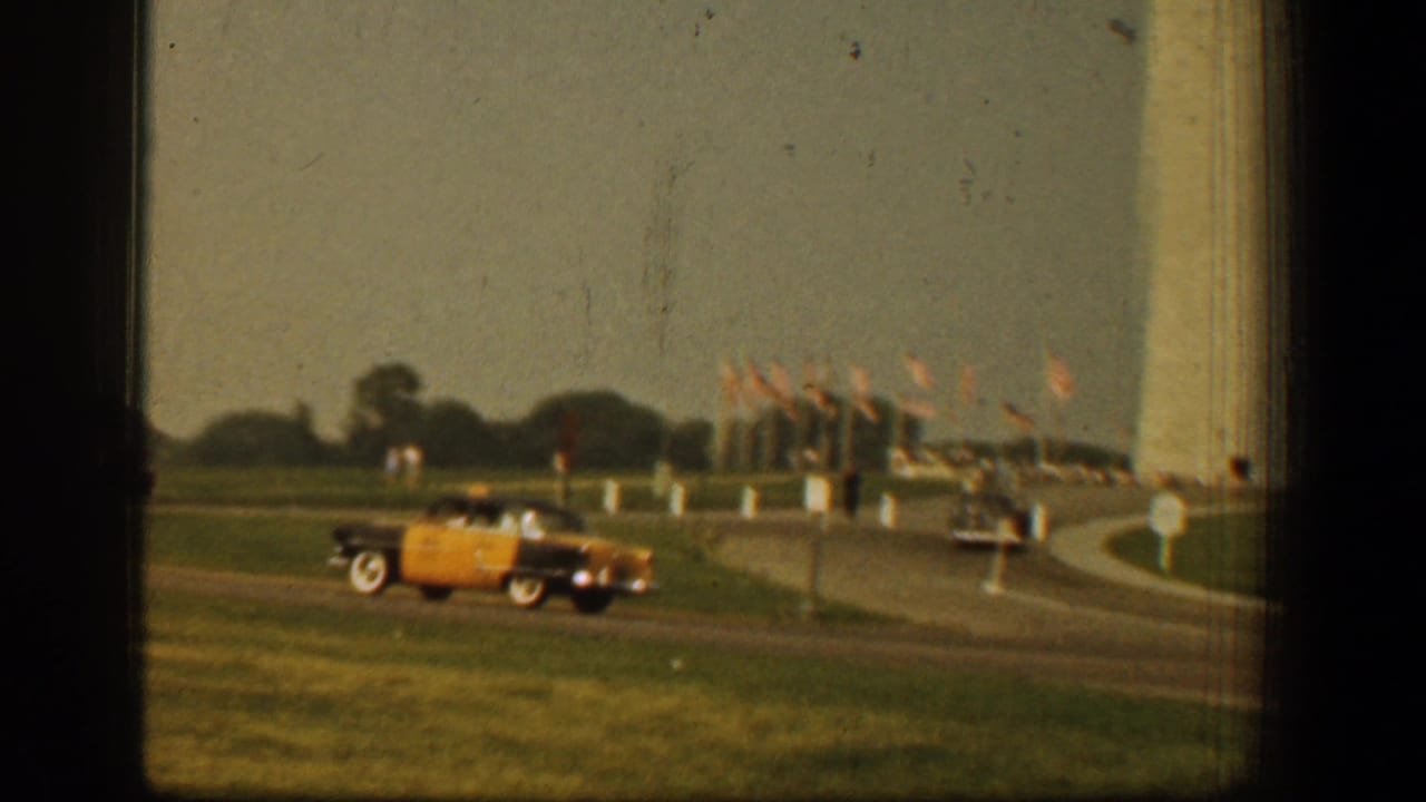 Classic car on grass near a curving road and flagpoles by the Washington Monument grounds.