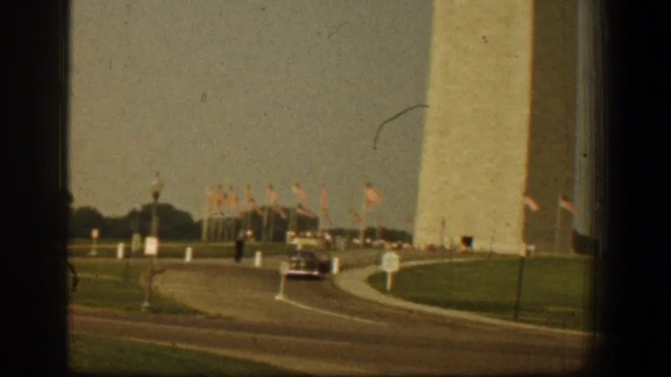 Washington Monument grounds in 1961 with a curving roadway, distant car, American flags, lawn, and soft 8mm exposure.