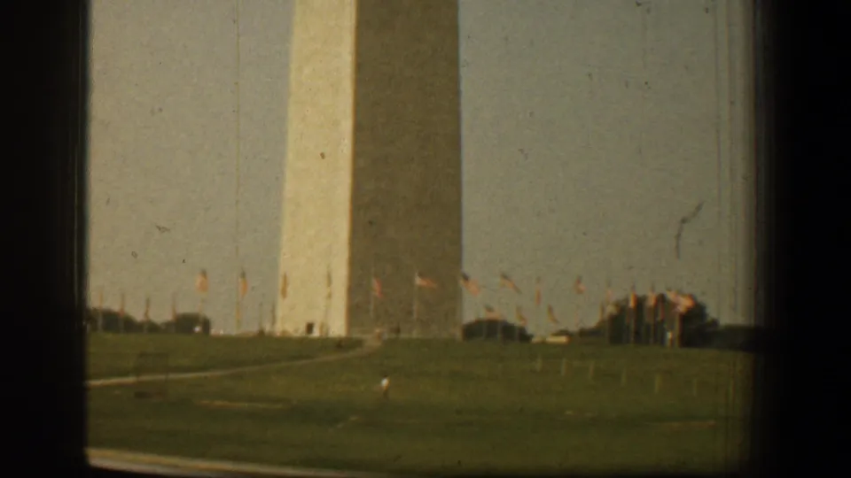 1961 Washington, DC home-movie frame of the Washington Monument with broad lawns, rows of American flags, and distant visitors.