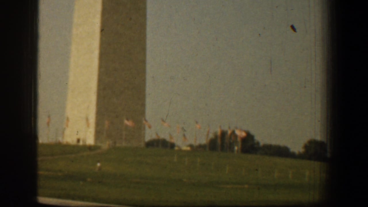1961 Washington, DC home-movie frame showing the Washington Monument across a grassy field with small flags and dark window-like edges.