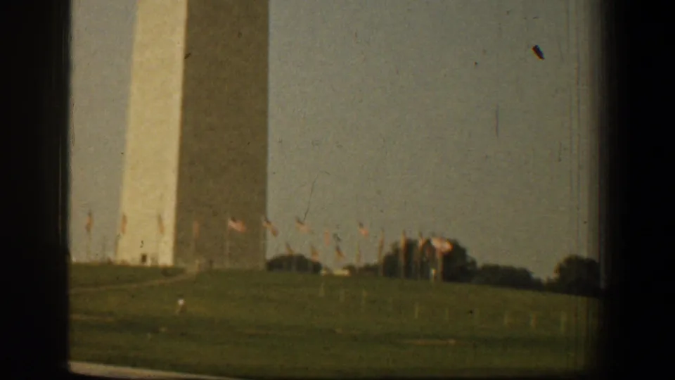 1961 Washington, DC home-movie frame showing the Washington Monument across a grassy field with small flags and dark window-like edges.