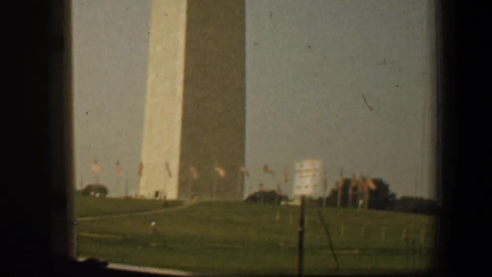 Washington, DC frame with the Washington Monument partly framed by dark edges, flags on the lawn, and a foreground sign.
