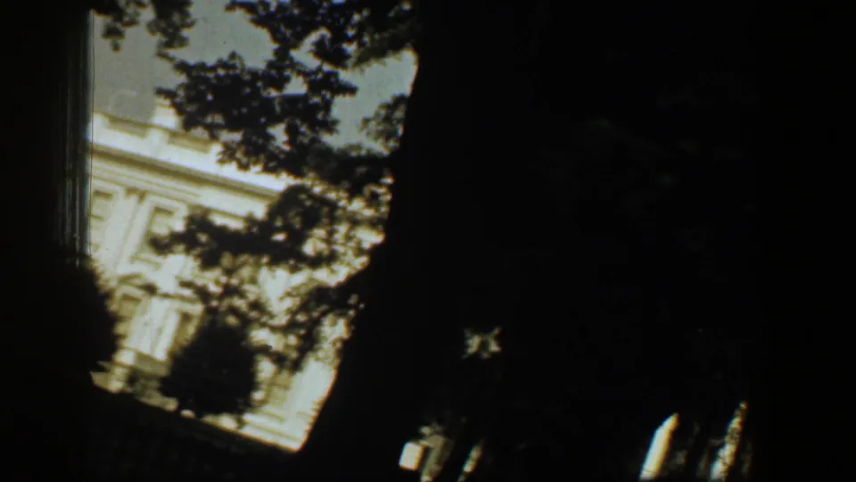 Tilted 1961 home-movie frame of the White House facade, partly blocked by tree branches and deep shadow.
