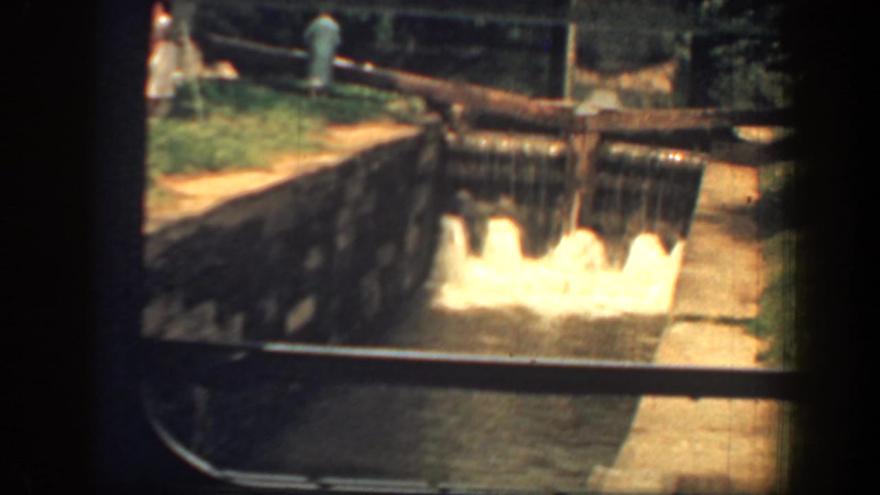 1961 8mm home-movie frame showing water pouring over a small spillway, with masonry sides and a rail or pipe above.