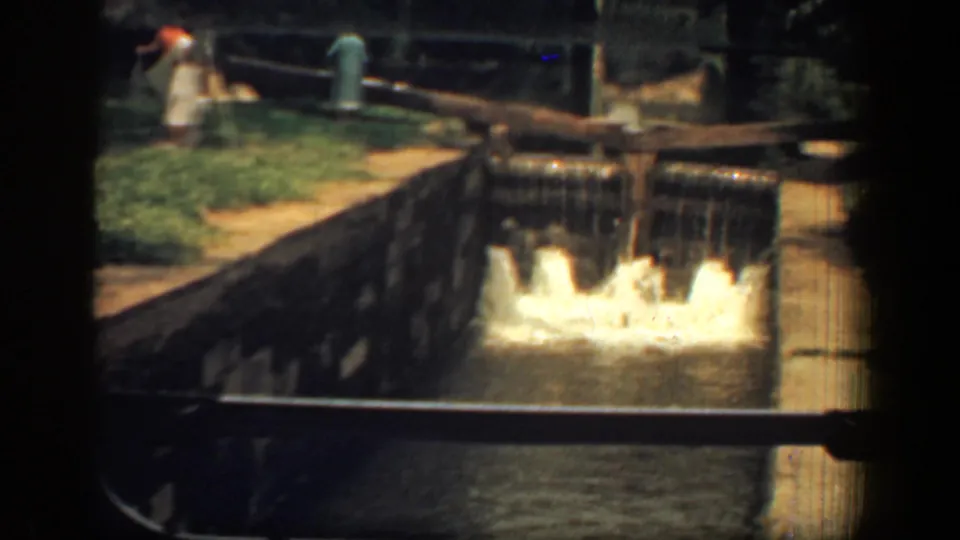 1961 8mm frame of a small dam or spillway with foaming water, masonry edges, and a person faintly visible near the upper left.