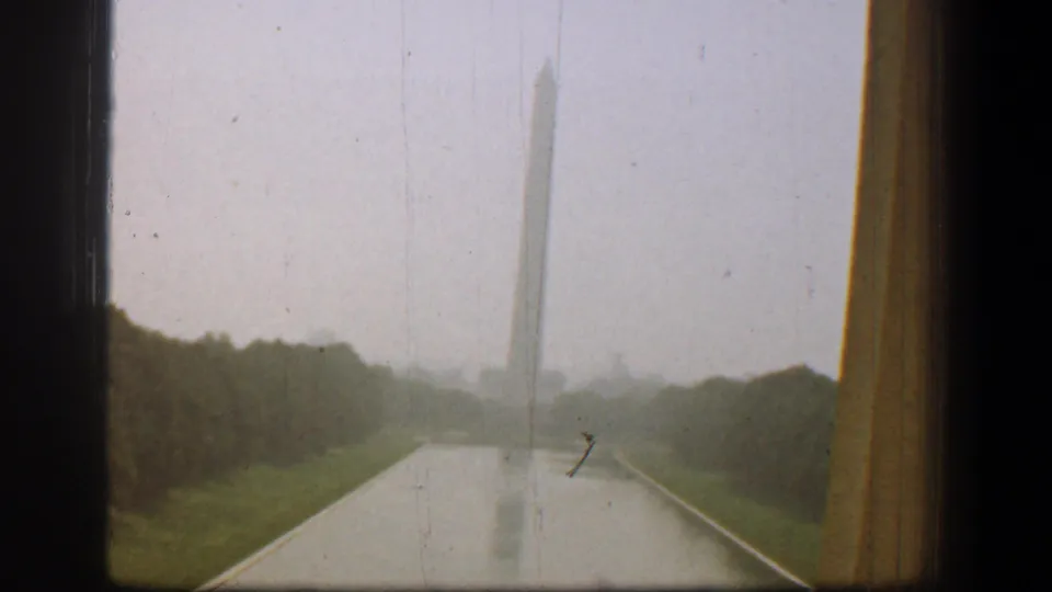 Rainy home-movie view of the Washington Monument from indoors or a vehicle, framed by a window and wet foreground surface.